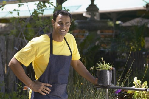 A man wearing a yellow t-shirt and a dark apron stands confidently outdoors in a residential garden, smiling at the camera. He is positioned next to a small round metal table that holds a black plastic pot containing a small plant with green foliage and purple flowers. The garden features well-maintained grass, edged by flower beds with various plants and shrubs, and is bordered by a wooden fence and dense hedges. In the background, there are trees, a shed, and garden structures, indicative of a typical backyard setting in Northolt, West London. The scene suggests an outdoor gardening activity, with natural daylight illuminating the lush greenery and colorful flowers, consistent with professional garden maintenance or landscaping work by Lawn Mowing Northolt, in a calm and well-kept outdoor environment.