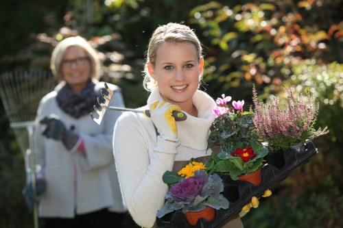 A young woman with blonde hair, wearing a white sweater and yellow gardening gloves, is standing in a well-maintained backyard garden during daytime, holding a black tray of flowering plants with colorful blooms and lush green foliage. Behind her, an older woman with glasses, a scarf, and a light jacket is smiling and holding a gardening fork, indicating involvement in garden maintenance activities. The garden features a neatly trimmed lawn, bordered by flower beds with various plants, and is illuminated by natural sunlight, creating a bright and welcoming outdoor environment suitable for gardening and landscaping services in Northolt. The mature trees and shrubbery in the background provide natural backdrop and privacy, reflecting a landscaped outdoor space that benefits from professional gardening care.