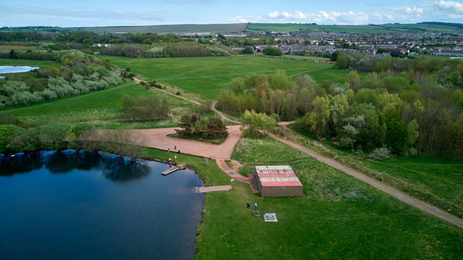 An aerial view of Northala Fields shows a landscaped park with well-maintained grassy lawns and pathways. The scene features a variety of trees and shrubs, some forming natural borders around open spa